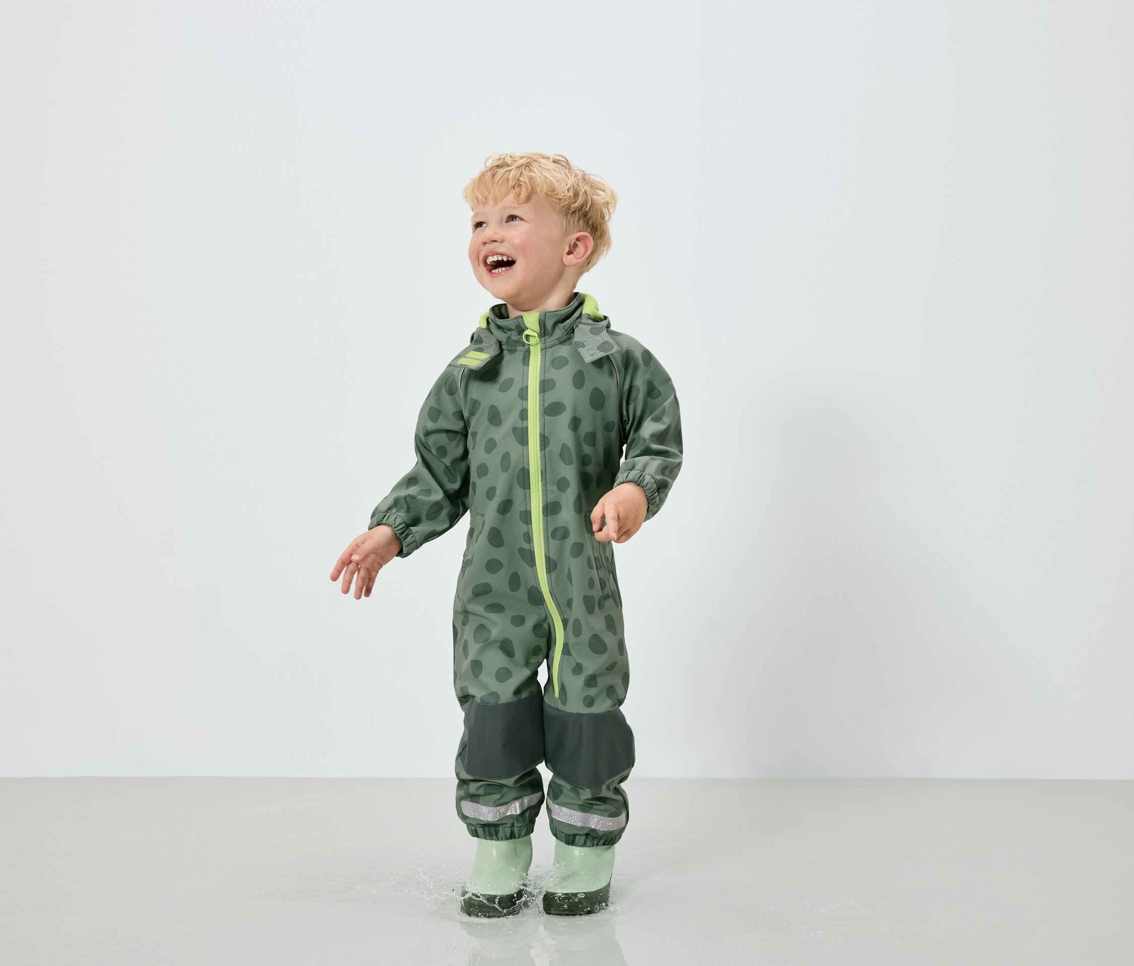 Ein Junge im grünen Overall mit Fleckenmuster und grünen Gummistiefeln springt in eine Pfütze.