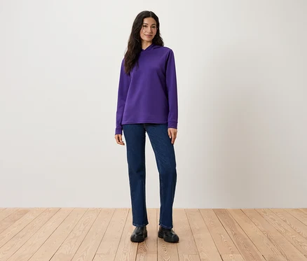 Eine Frau steht und trägt einen violetten Kapuzenpullover, Jeans und schwarze Stiefel.