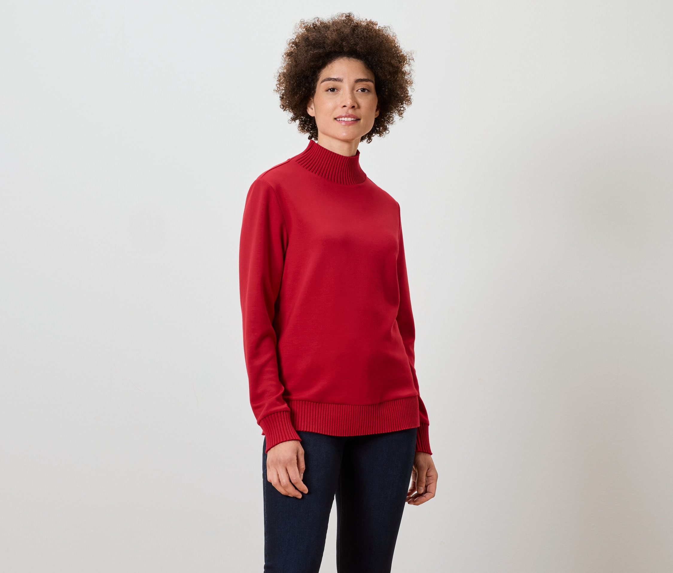 Frau mit lockigem Haar steht und trägt einen roten Rollkragenpullover und dunkelblaue Jeans.