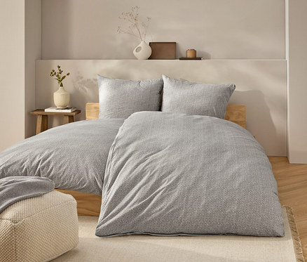 Bett mit grauer Bettwäsche und zwei Kissen. Auf dem Bett liegt eine graue Decke. Neben dem Bett steht ein beiger Pouf.