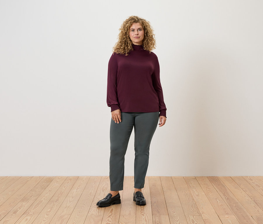 Eine Frau steht auf einem Holzboden und trägt eine dunkelgrüne Stretchhose und ein weinrotes Langarmshirt.