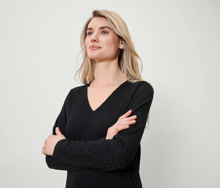 Blonde Frau mit verschränkten Armen trägt ein schwarzes Langarmshirt mit V-Ausschnitt.