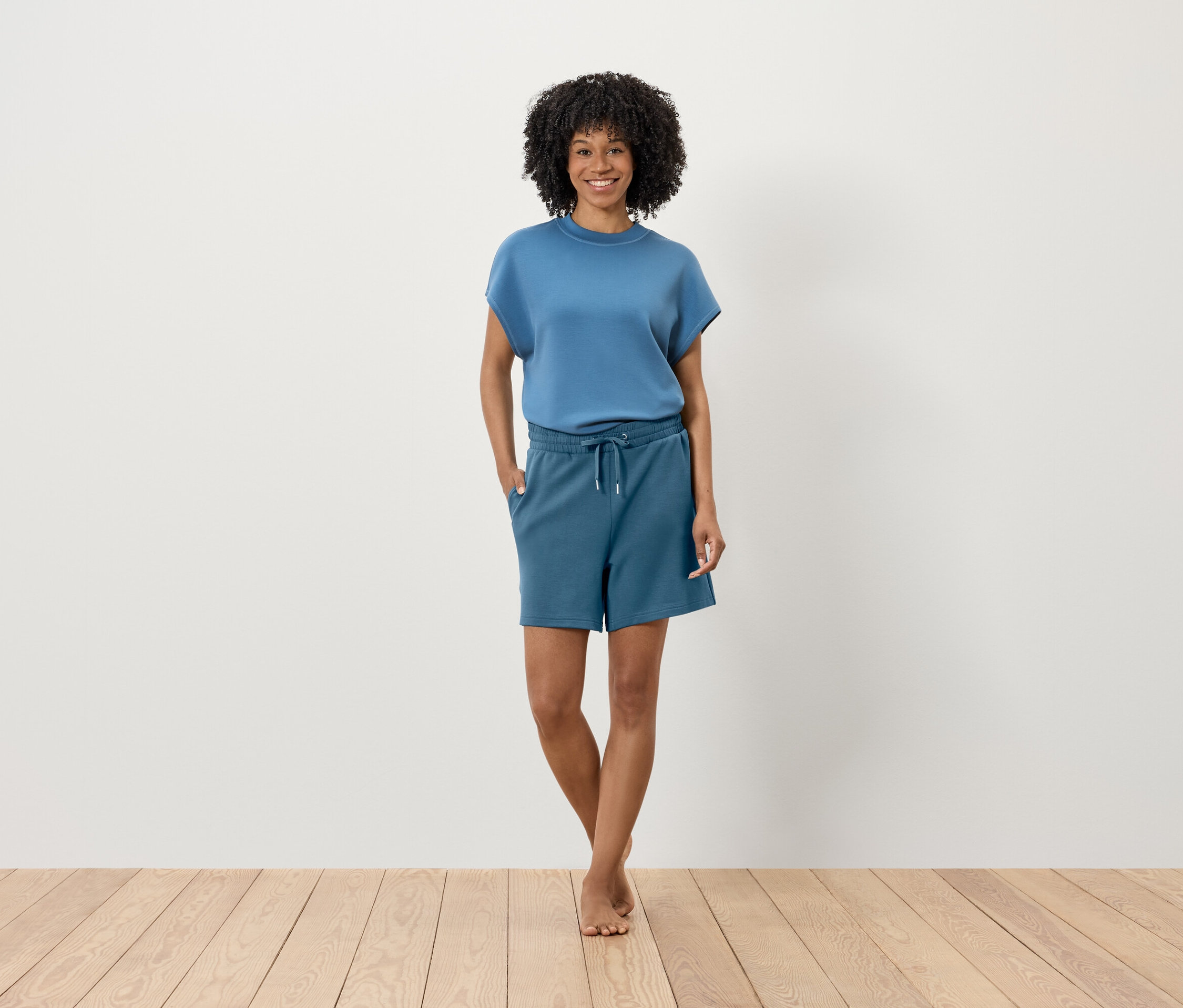 Eine Frau mit Afro-Frisur steht auf einem Holzboden vor einer weißen Wand. Sie trägt ein blaues T-Shirt und Shorts.