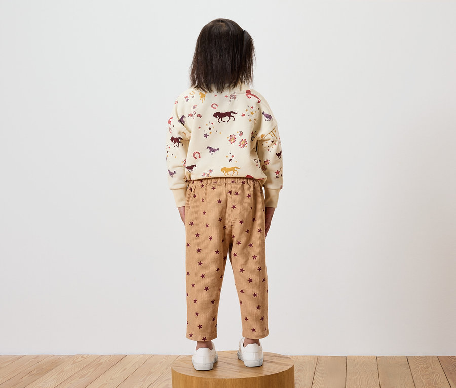 Ein Mädchen steht mit dem Rücken zur Kamera und trägt ein cremefarbenes Sweatshirt mit Pferdeaufdruck und braune Hosen mit Sternen.