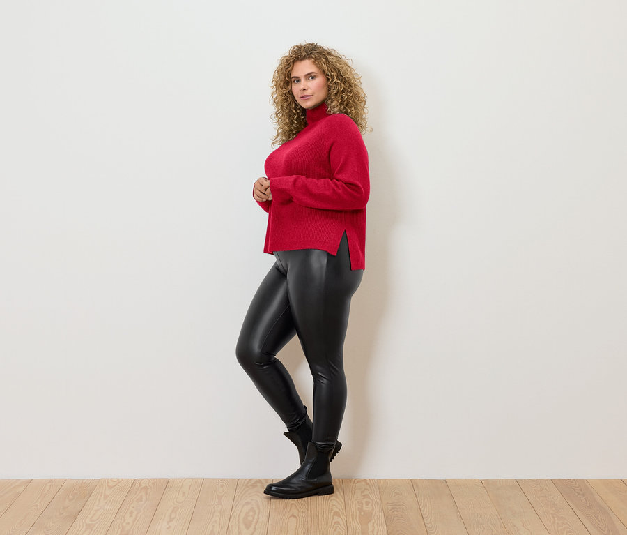 Frau mit lockigem Haar posiert seitlich und trägt einen roten Rollkragenpullover, schwarze Leggings und schwarze Stiefel.