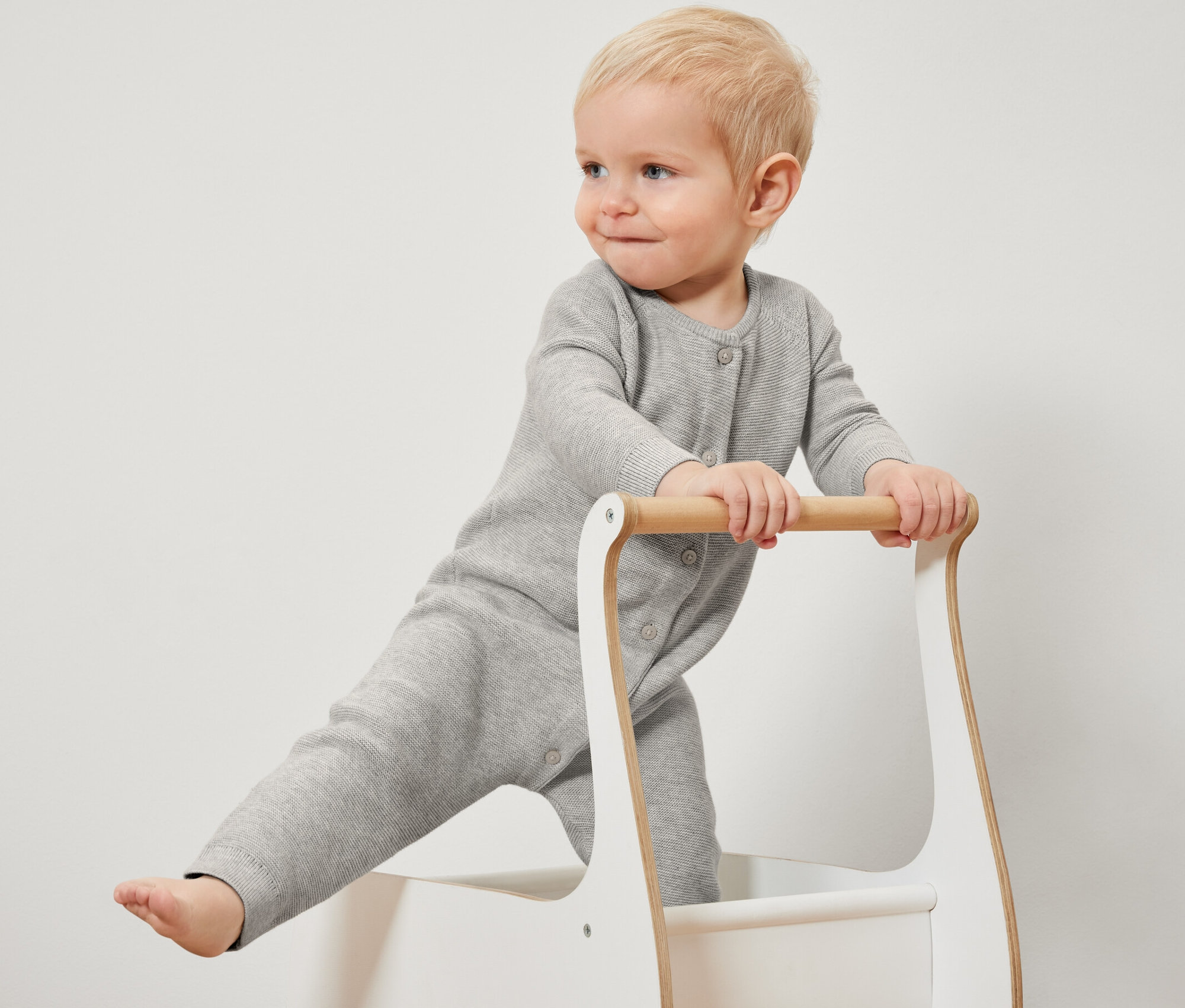 Ein blondes Kleinkind steht in einer Lauflernhilfe und trägt einen grauen Strickoverall.
