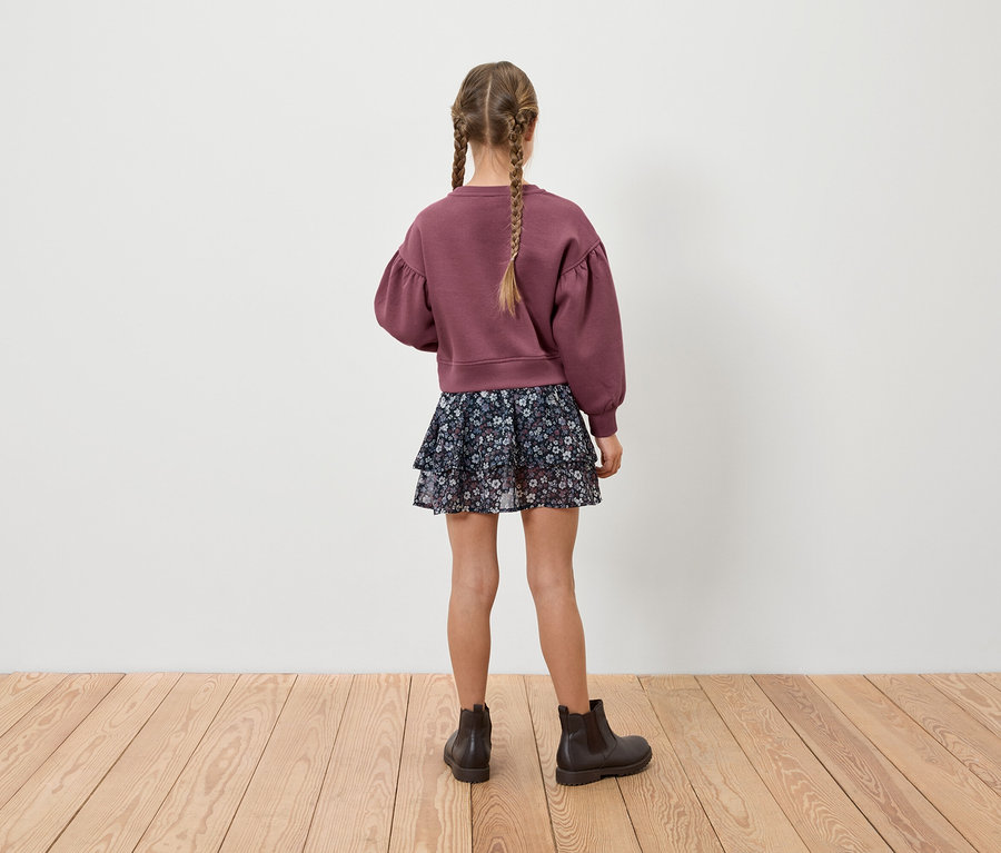 Mädchen mit Zöpfen steht mit dem Rücken zur Kamera, trägt ein weinrotes Sweatshirt, einen Blumenrock und braune Stiefel.