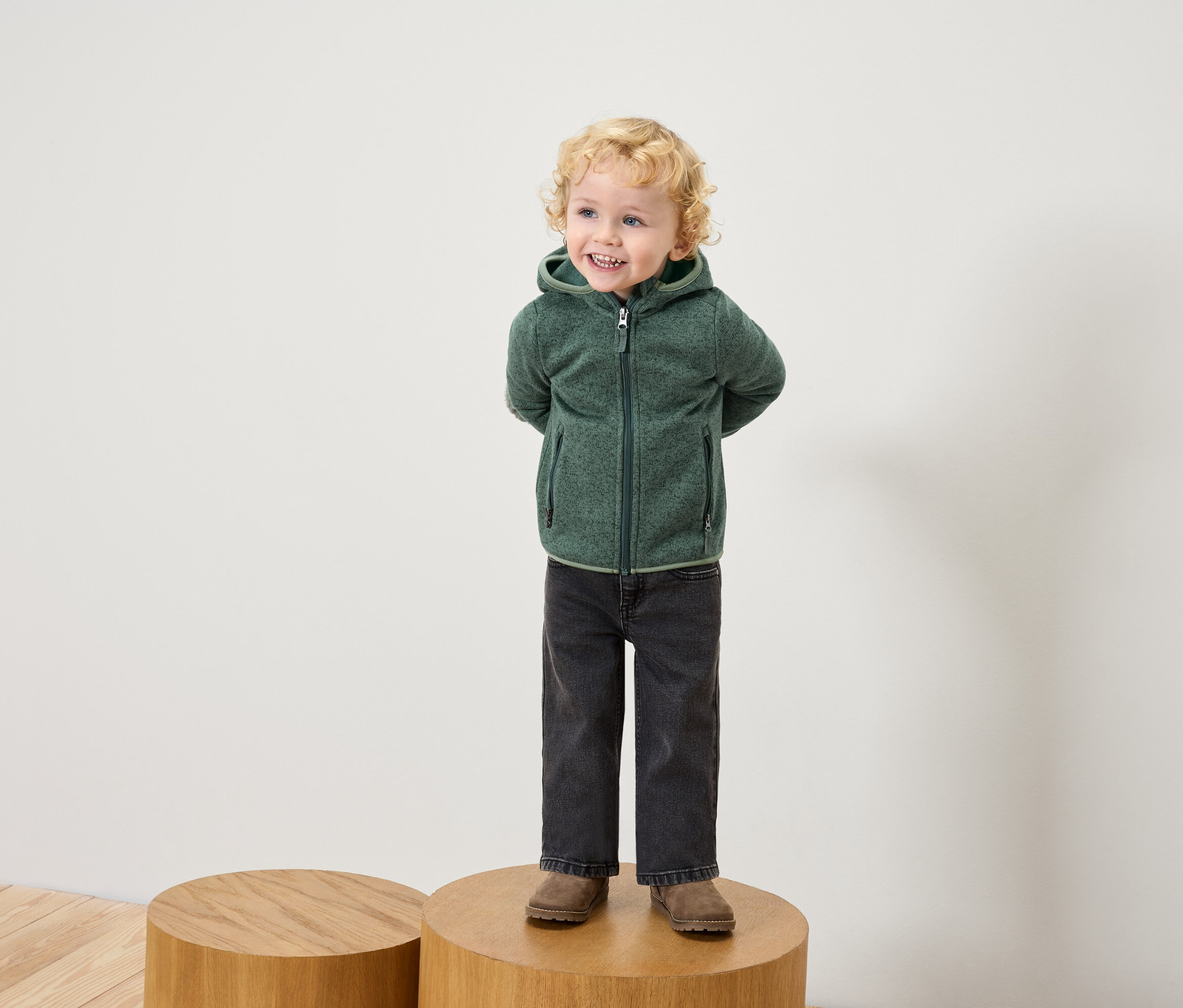 Ein Junge mit blond gelocktem Haar steht auf einem Holzsockel und trägt eine grüne Fleecejacke mit Kapuze, schwarze Jeans und braune Schuhe.