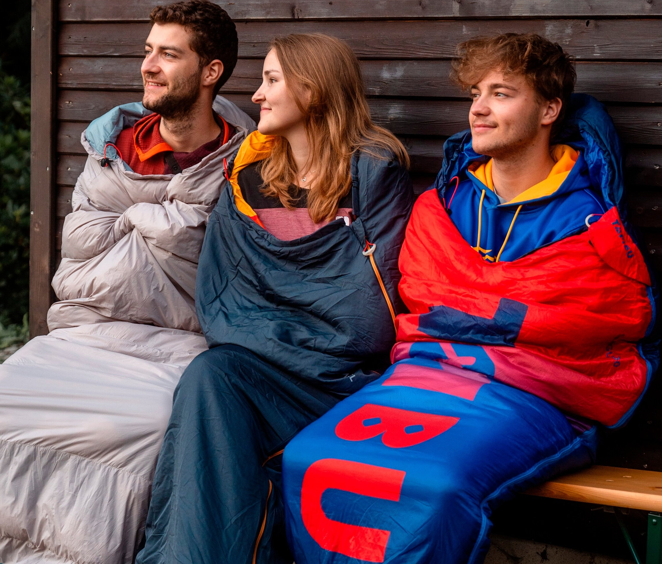Drei Personen sitzen nebeneinander auf einer Bank, eingewickelt in Tambu Mumienschlafsäcke »SANGU 600«.