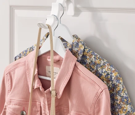 Rosa Jeansjacke und geblümtes Hemd hängen an einem weißen Kleiderbügel, der an einem weißen Türhänger befestigt ist.