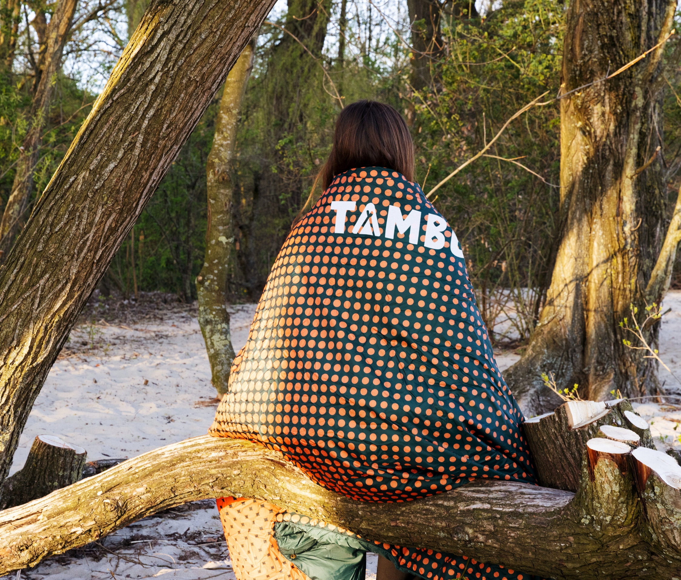 Frau sitzt mit Tambu Campingdecke »KAMBALA« auf Baumstamm im Wald.