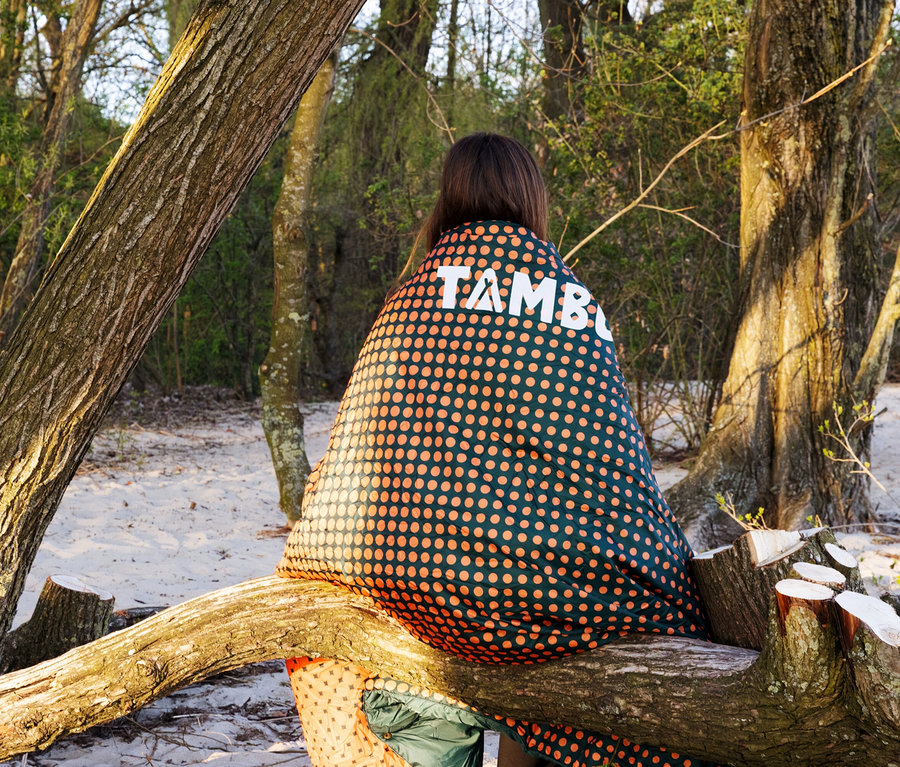 Frau sitzt mit Tambu Campingdecke »KAMBALA« auf Baumstamm im Wald.