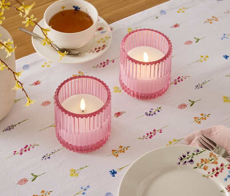 Auf einem Tisch mit Blumenmuster stehen zwei rosa Kerzenhalter, eine Tasse Tee und ein Teller.