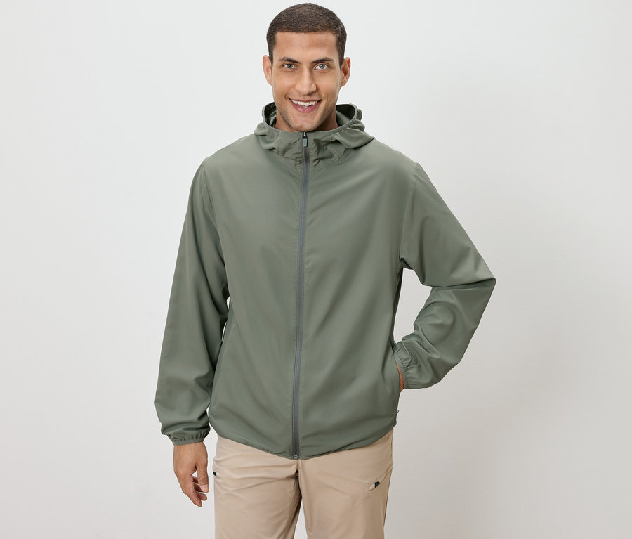 Mann mit olivgrüner Jacke und beiger Hose, eine Hand in der Tasche, vor einem weißen Hintergrund.