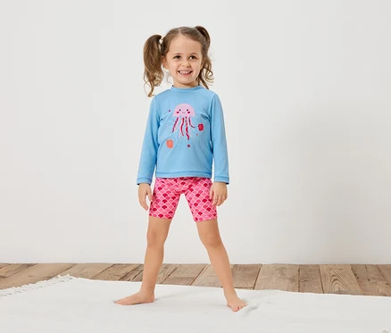 Mädchen mit Zöpfen trägt ein hellblaues Langarmshirt mit einer rosa Qualle und rosafarbene Shorts mit Schuppenmuster.