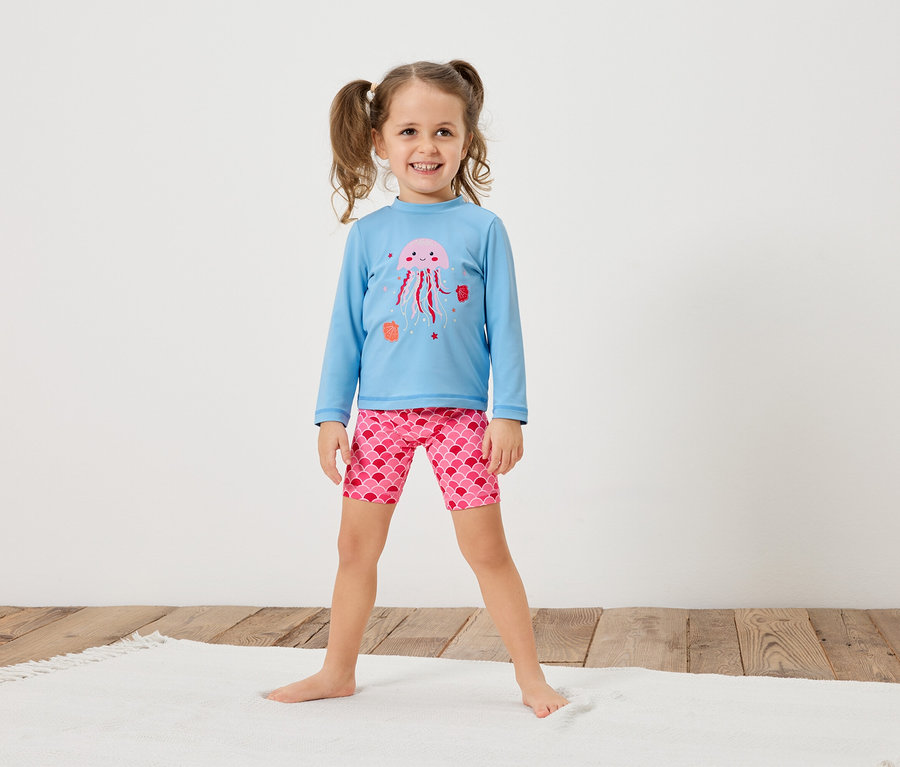 Mädchen mit Zöpfen trägt ein hellblaues Langarmshirt mit einer rosa Qualle und rosafarbene Shorts mit Schuppenmuster.