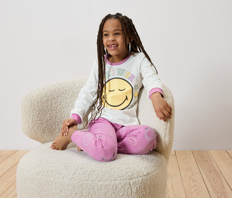 Lächelndes Mädchen mit Zöpfen sitzt barfuß in einem Sessel und trägt einen weißen Schlafanzug mit Smiley-Motiv.