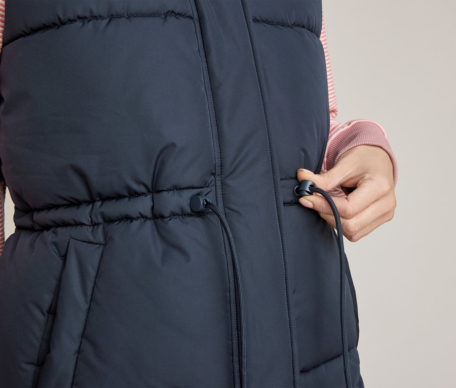 Nahaufnahme einer Person in einer dunkelblauen Steppweste über einem rosa gestreiften Hemd, die den Kordelzug an der Taille der Weste mit der rechten Hand verstellt.
