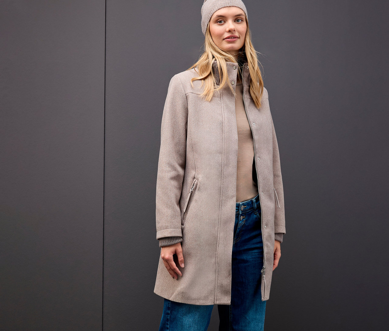 Blonde Frau trägt einen Street One Kurzmantel, eine Mütze, einen Rollkragenpullover und Jeans vor einem grauen Hintergrund.