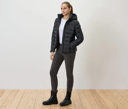 Frau trägt eine bedruckte Steppjacke mit Kapuze, Jeans und schwarze Stiefel.