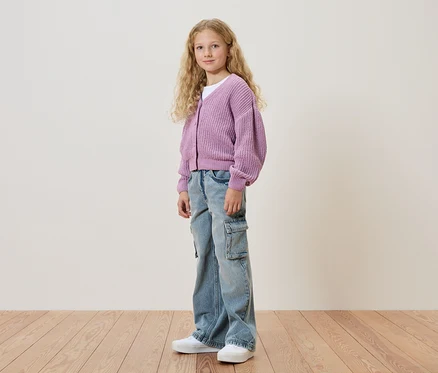 Mädchen mit blonden, lockigen Haaren, trägt eine lila Strickjacke, eine Jeanshose und weiße Schuhe.