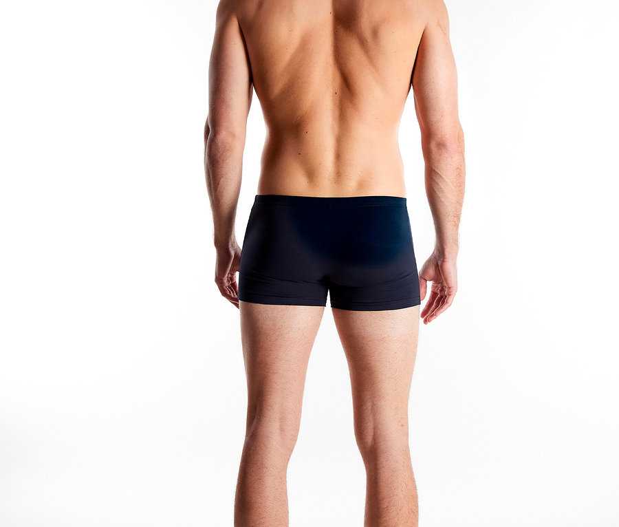 Rückansicht eines Mannes mit nacktem Oberkörper und dunklen Boxershorts vor einem weißen Hintergrund.