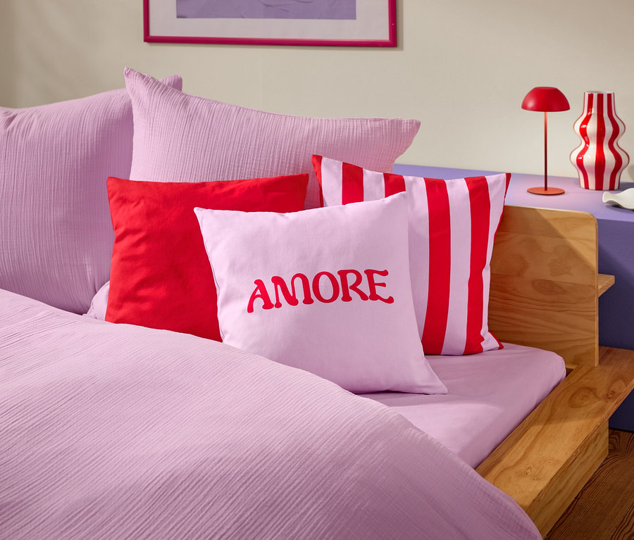 Nahaufnahme eines Bettes mit rosa Bettwäsche und Kissen. Ein Kissen hat das Wort AMORE in roter Schrift.