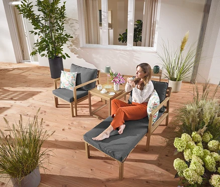 Eine Frau sitzt mit einem Getränk auf einem ausziehbaren Gartensessel. Eine LED-Akku-Outdoor-Tischleuchte in Salbei steht auf einem Tisch neben ihr. Im Hintergrund sind 2 Outdoor-Kissen mit floralem Muster zu sehen.