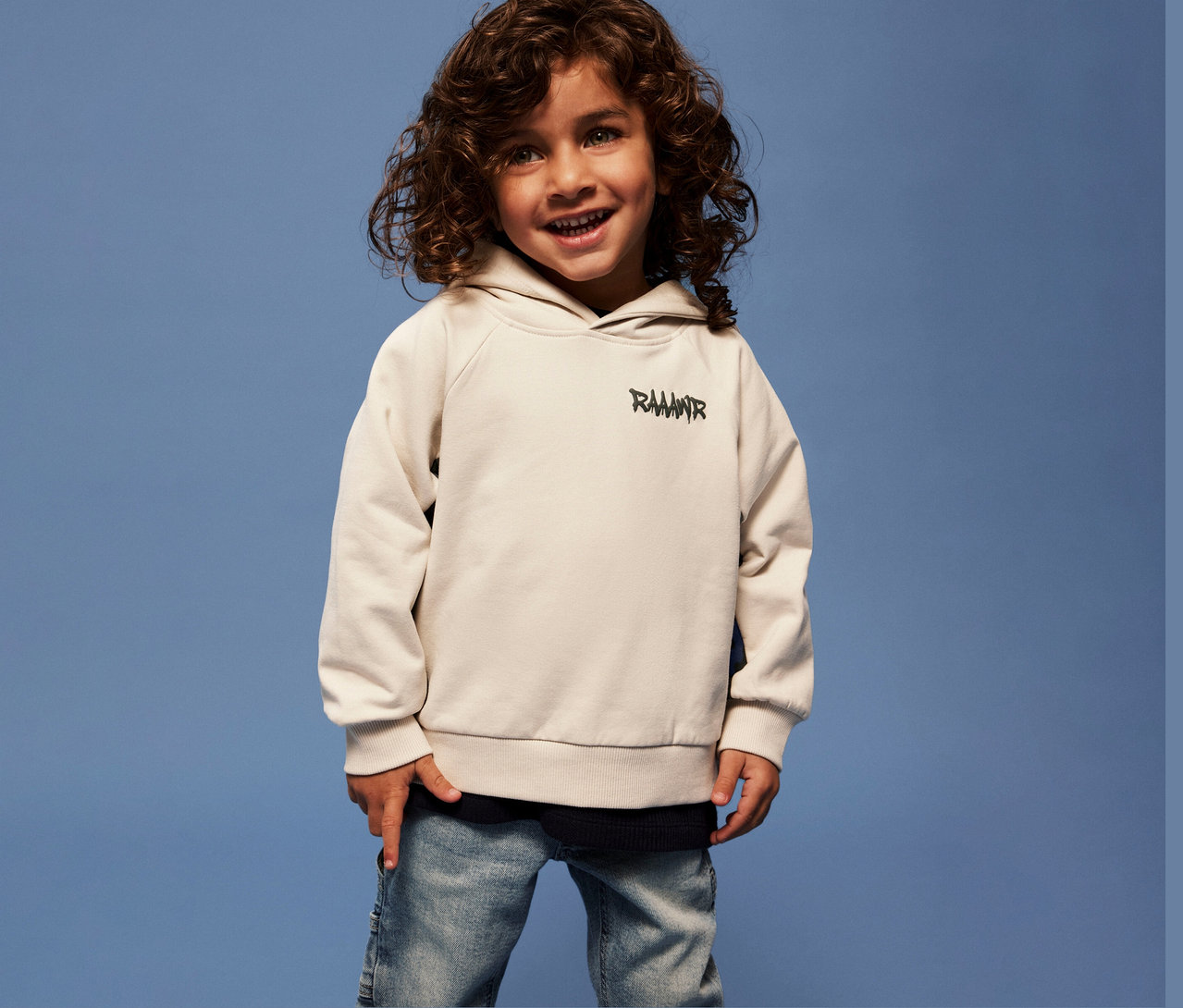 Lächelnder Junge mit lockigem Haar trägt ein NAME IT Kinder-Sweatshirt mit 3D-Dino-Applikation und Jeans vor blauem Hintergrund.