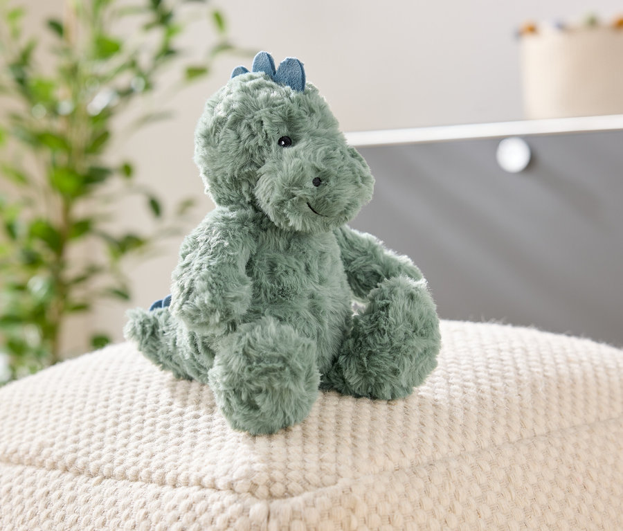Ein grüner, plüschiger Dinosaurier sitzt auf einem weißen, strukturierten Hocker. Im Hintergrund sind eine Pflanze und ein Schrank zu sehen.