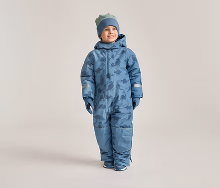 Ein lächelnder Junge trägt einen blauen Schneeanzug mit Dinosaurier-Muster, passende Handschuhe und eine Mütze vor einem hellen Hintergrund.