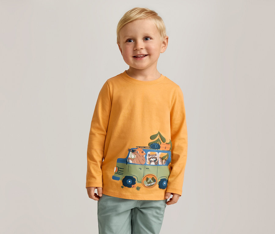 Lächelnder Junge mit blonden Haaren trägt ein orangefarbenes Langarmshirt mit einem grünen Autoaufdruck.