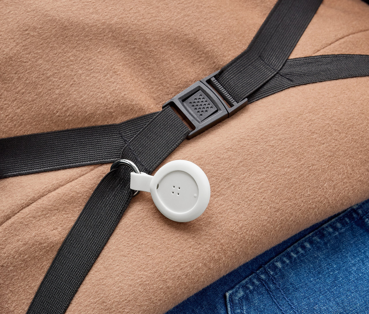 Ein weißes, rundes Gerät mit drei Punkten ist an einem schwarzen Gurt mit einem Metallring befestigt. Der Gurt mit einer schwarzen Kunststoffschnalle liegt über hellbraunem Stoff und blauem Jeansstoff.