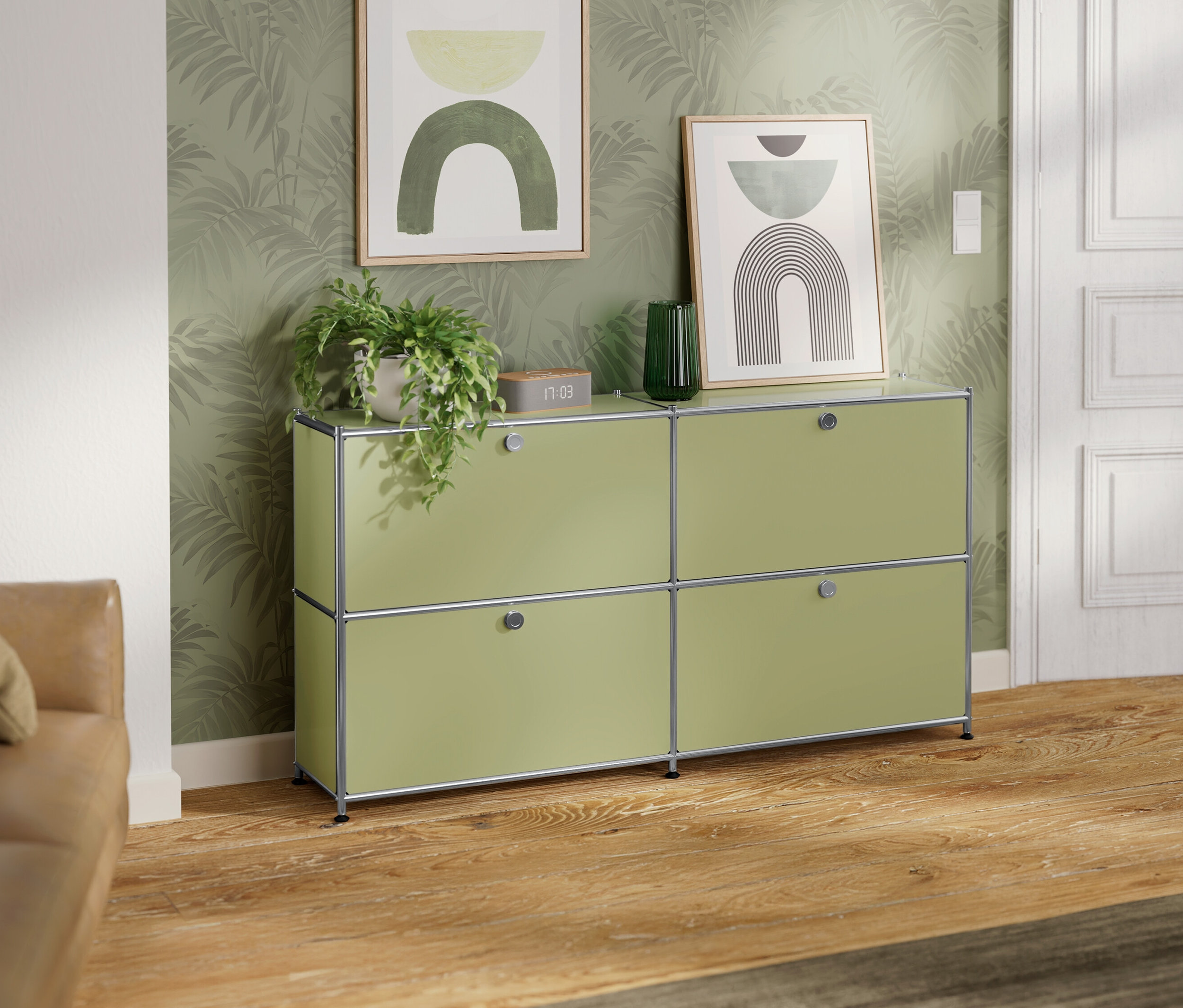 Ein hellgrünes Sideboard mit vier Fächern steht vor einer Wand mit Palmenmuster. Auf dem Sideboard stehen eine Pflanze, eine Uhr und eine grüne Vase. An der Wand hängen zwei gerahmte Bilder.