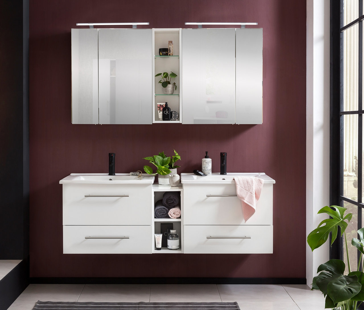 Modernes Badezimmer mit doppelter Waschtischkombination und Spiegelschrank »Dorina« mit Licht. Der weiße Waschtisch hat zwei Waschbecken, schwarze Armaturen und Schubladen. Der Spiegelschrank hat Spiegeltüren und offene Regale. Die Wand ist weinrot.