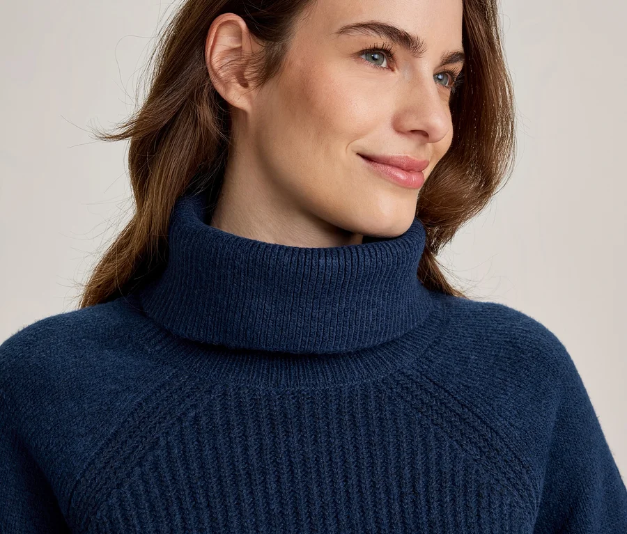 Nahaufnahme einer Frau in einem marineblauen Rollkragenpullover, die mit einem leichten Lächeln nach rechts blickt. Ihr braunes Haar umrahmt ihr Gesicht, der Pullover hat ein Rippenmuster.