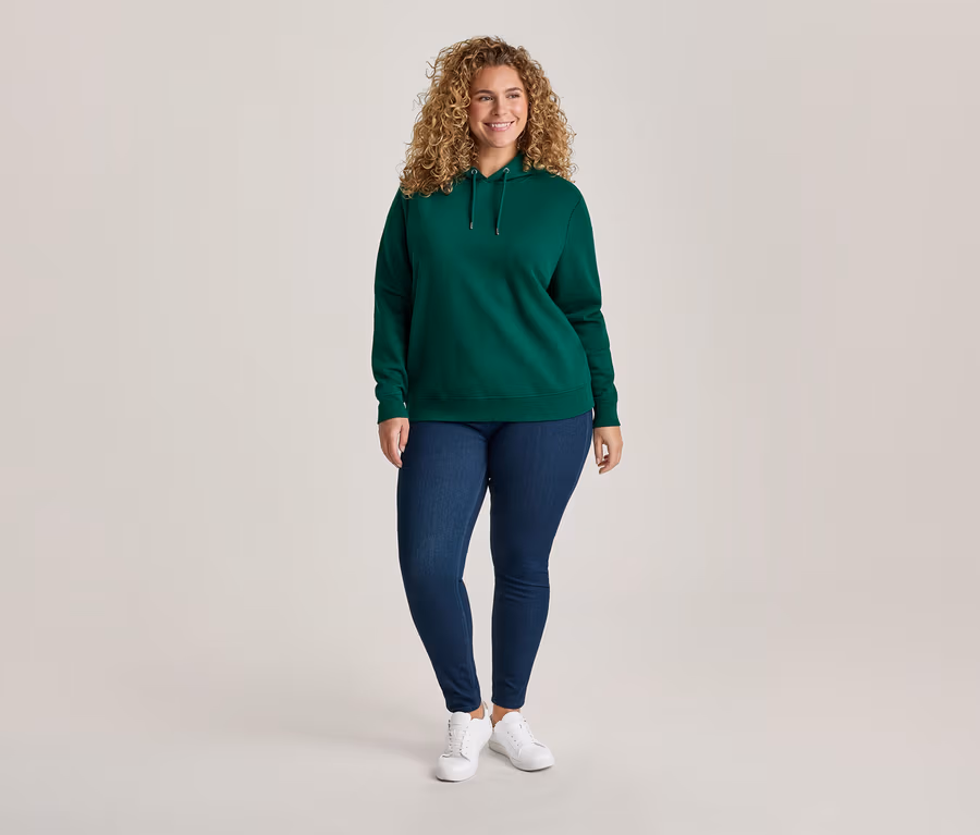 Lächelnde Frau mit lockigem Haar trägt einen grünen Hoodie, blaue Jeans und weiße Turnschuhe vor hellem Hintergrund.