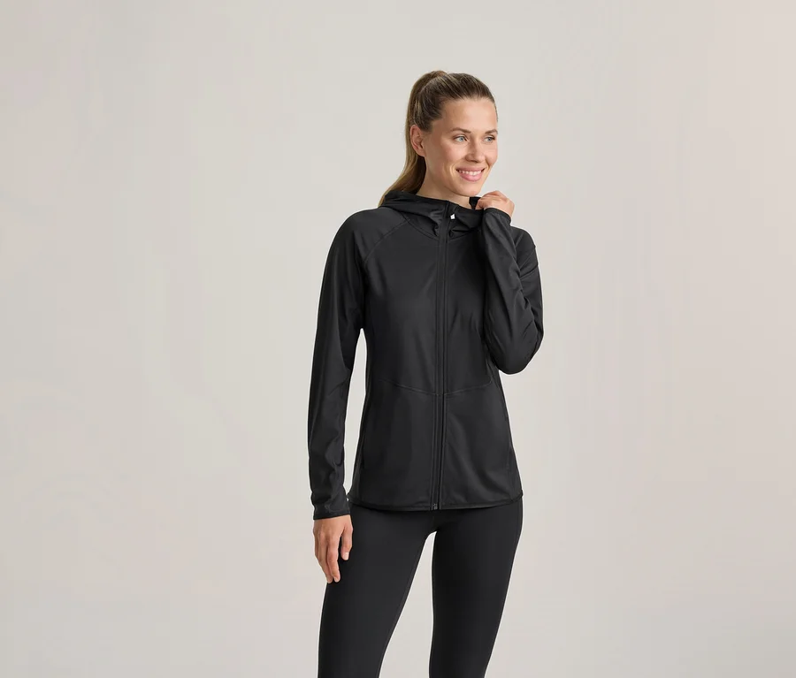 Eine Frau in einer schwarzen Kapuzenjacke und schwarzen Leggings, lächelt und blickt nach rechts, ihre linke Hand an der Kapuze. Ihr Haar ist zu einem Pferdeschwanz gebunden.