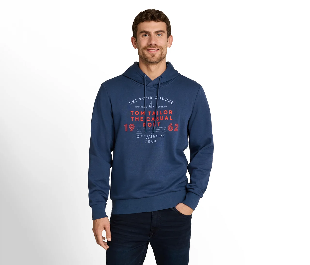 Mann mit dunklem Haar und Bartstoppeln trägt einen marineblauen Hoodie mit 'TOM TAILOR' Grafikdruck und dunkle Jeans, steht mit einer Hand in der Tasche.