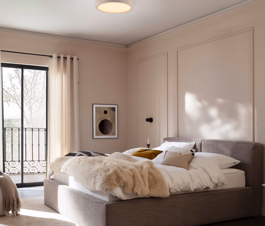 Ein Schlafzimmer mit einem großen, gepolsterten Bett mit weißer Bettwäsche und einer flauschigen, beigen Decke. Über dem Bett hängt eine runde Deckenleuchte, und an der Wand befindet sich ein abstraktes Bild. Links führt eine Glastür zu einem Balkon.