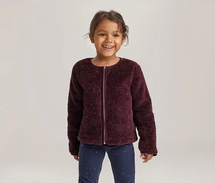 Lächelndes kleines Mädchen in einer bordeauxroten, flauschigen Jacke mit Reißverschluss und dunkelblauen gemusterten Hosen.