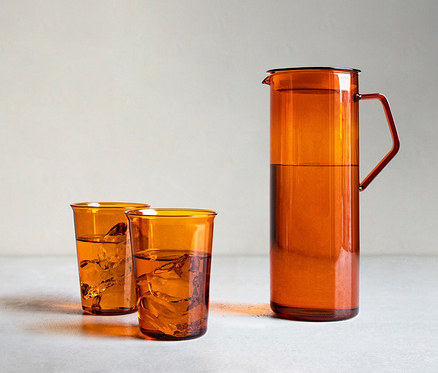 Bernsteinfarbener KINTO Krug Cast Amber mit schwarzem Deckel und rotem Griff, sowie zwei passende Gläser, alle mit Wasser und Eiswürfeln gefüllt.
