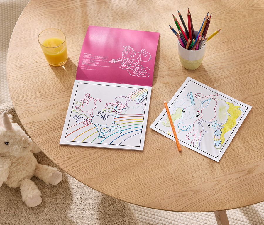 Ein pinkes Einhorn-Malbuch liegt offen auf einem Holztisch neben einer teilweise ausgemalten Einhornseite, einem Becher mit Buntstiften und einem Glas Orangensaft. Ein flauschiges weißes Einhorn-Stofftier sitzt auf einem beigen Sessel neben dem Tisch.