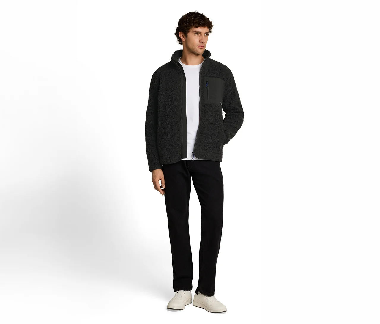 Stehender Mann in einer dunkelgrünen Fleecejacke mit Reißverschluss und Brusttasche, weißem T-Shirt, schwarzer Hose und weißen Turnschuhen.