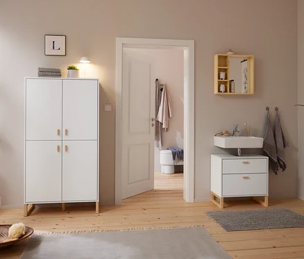 Helles Badezimmer mit Holzboden und hellgrauen Wänden. Links steht ein weißer Schrank mit einer weißen LED-Akku-Tischleuchte und einer Pflanze. Rechts hängt ein Spiegel aus massivem Bambus mit Ablagen über einem Waschbecken mit Unterschrank. Eine offene Tür führt in einen Nebenraum.