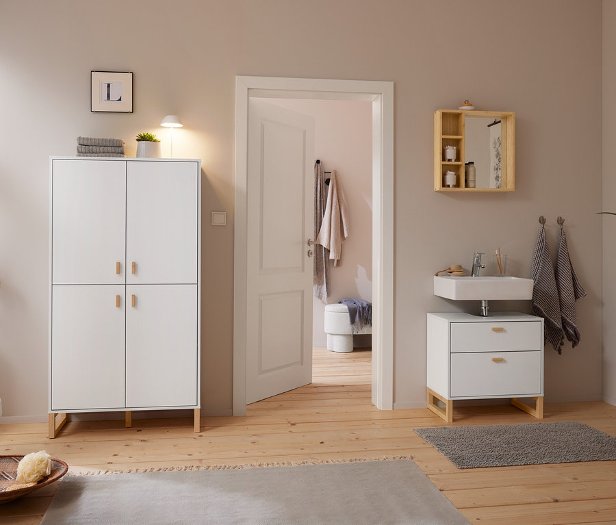 Helles Badezimmer mit Holzboden und hellgrauen Wänden. Links steht ein weißer Schrank mit einer weißen LED-Akku-Tischleuchte und einer Pflanze. Rechts hängt ein Spiegel aus massivem Bambus mit Ablagen über einem Waschbecken mit Unterschrank. Eine offene Tür führt in einen Nebenraum.