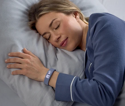 Eine Frau schläft friedlich auf der Seite auf einem grauen Kissen, blaue Pyjamas tragend. Ein grauer Fitness-Tracker an ihrem linken Handgelenk zeigt "58" und "80" an.