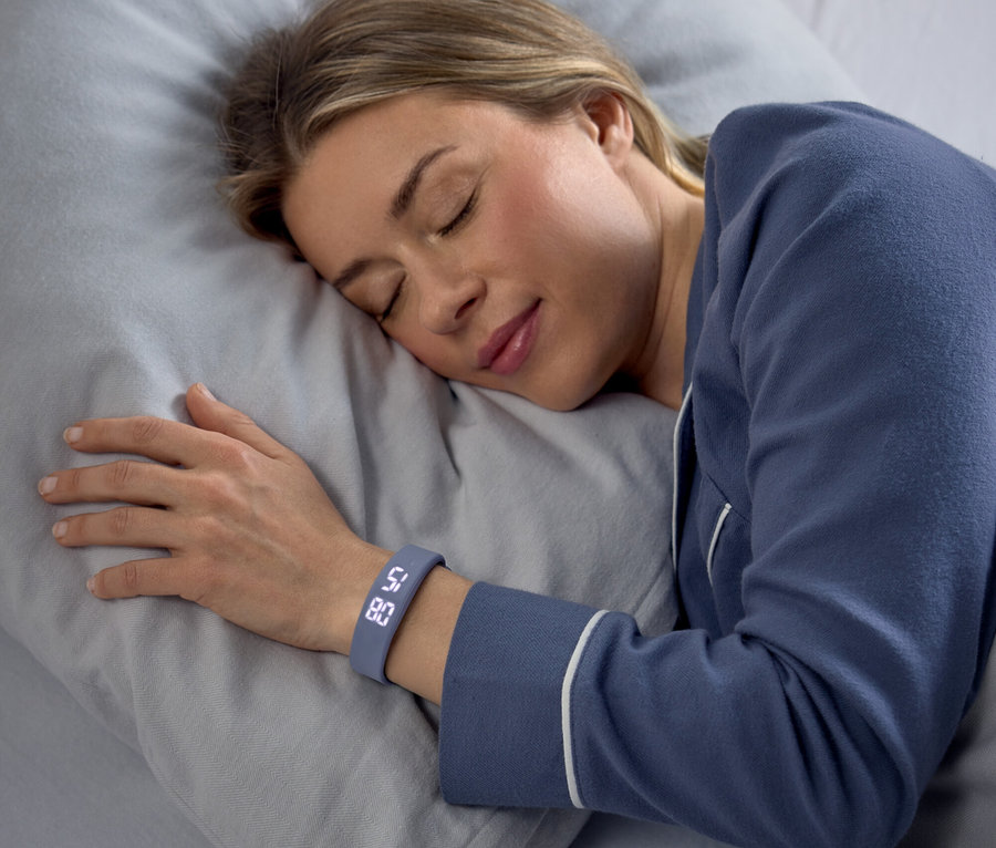 Eine Frau schläft friedlich auf der Seite auf einem grauen Kissen, blaue Pyjamas tragend. Ein grauer Fitness-Tracker an ihrem linken Handgelenk zeigt "58" und "80" an.
