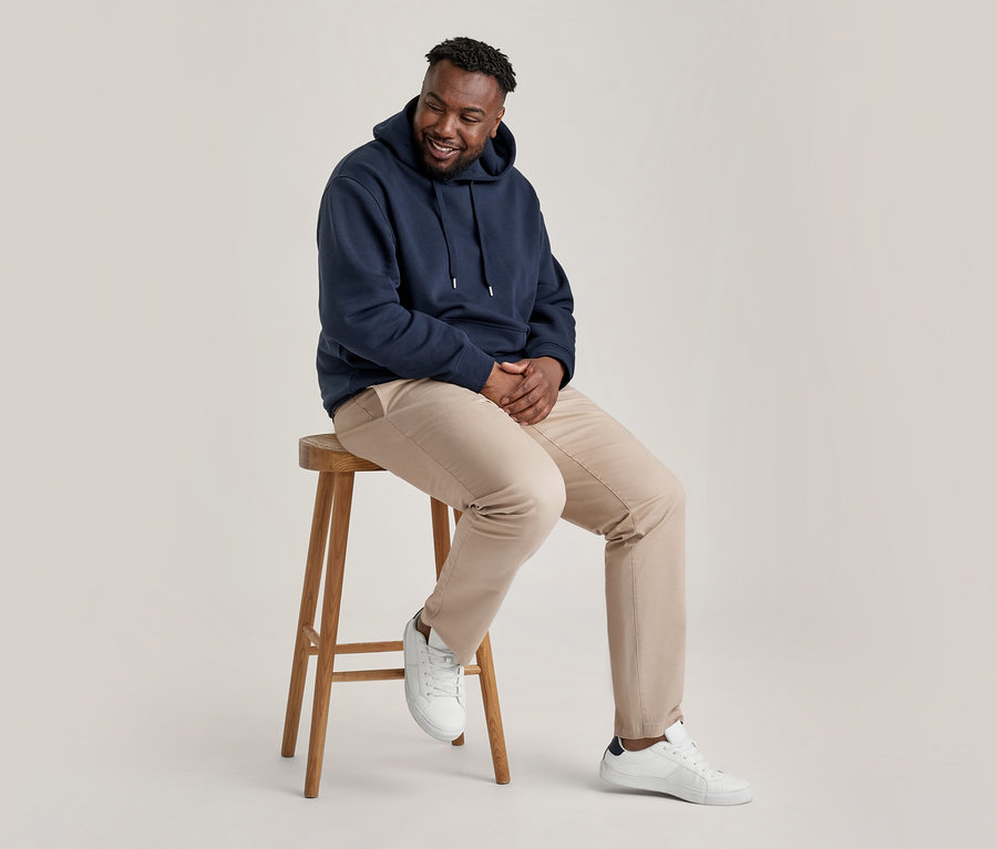 Ein lächelnder Mann mit Bart sitzt auf einem Holzschemel und trägt einen marineblauen Hoodie, eine beige Chino und weiße Sneaker.