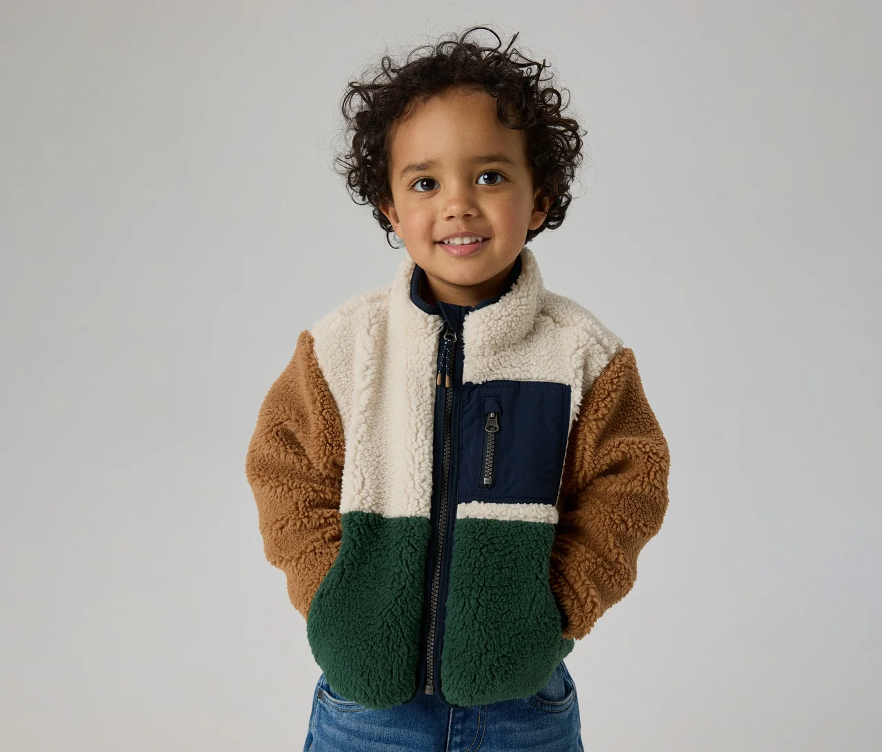 Lächelndes Kind mit lockigem Haar trägt eine Color-Block-Fleecejacke in Creme, Braun, Dunkelgrün und mit einer marineblauen Brusttasche.